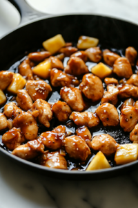 A black cast-iron skillet on the white marble cooktop, with diced boneless chicken thighs sizzling in hot avocado oil. The chicken is turning golden brown as steam rises from the pan.
