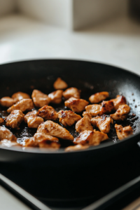 A close-up shot of a black skillet on the white marble cooktop, sizzling with marinated chicken pieces. The chicken is golden brown and slightly crispy on the edges as it cooks over medium-high heat, releasing rich, savory aromas.