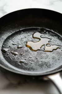 A close-up shot of a black skillet on a white marble cooktop, preheated and lightly greased with oil. The marinated chicken sizzles as it cooks, forming a golden, slightly charred crust on the surface while the aroma of smoky spices fills the air.