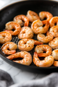 A black skillet on a white marble cooktop with plump shrimp sizzling in the oil. The shrimp are turning pink and opaque, seasoned with salt and pepper, creating a flavorful base for the dish.