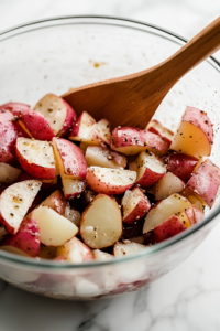 A close-up of a large glass mixing bowl on a white marble cooktop, filled with bite-sized chunks of cooled red potatoes. A wooden spoon rests on the edge of the bowl, ready for mixing.