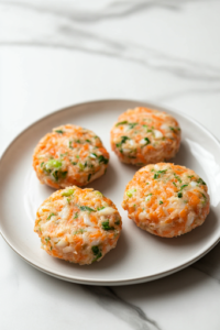 Four evenly shaped crab cake patties resting on a plate over a white marble cooktop. The patties are uniform in size and are set to firm up before cooking, ensuring they hold their shape during frying.