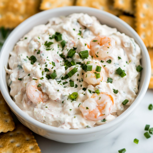 A close-up shot of a chilled serving bowl filled with creamy crab and shrimp dip on the white marble cooktop. The dip is garnished with extra chopped green onions and is surrounded by an assortment of crackers and toasted bread.A close-up shot of a chilled serving bowl filled with creamy crab and shrimp dip on the white marble cooktop. The dip is garnished with extra chopped green onions and is surrounded by an assortment of crackers and toasted bread.