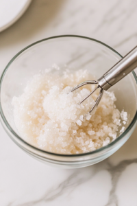 A close-up shot of a separate glass mixing bowl on the white marble cooktop, where softened butter and granulated sugar are being beaten into a light and creamy consistency. A hand mixer is partially visible, blending the ingredients smoothly for a soft and fluffy loaf texture.