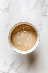 A stand mixer bowl on the white marble cooktop, where softened butter and granulated sugar are being creamed together into a smooth, fluffy mixture. A spatula rests on the side of the bowl, ready to scrape down the sides for even mixing.