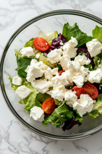 A close-up view of a hand crumbling fresh feta cheese over a colorful mixed salad in a glass bowl on a white marble cooktop. The creamy white cheese adds texture and a delicious contrast to the crisp vegetables.