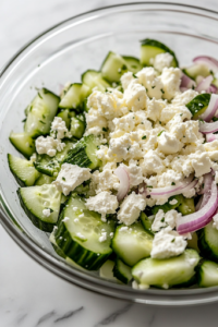 A close-up view of a hand crumbling fresh feta cheese over a glass bowl filled with cucumber, red onion, and avocado. The creamy white cheese adds a rich texture to the colorful mixture, enhancing the dish’s overall appeal.