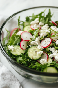 A close-up view of a glass mixing bowl on a white marble cooktop with creamy goat cheese being crumbled over the salad. The soft white cheese contrasts beautifully with the vibrant greens and crisp vegetables, adding a rich, tangy flavor.