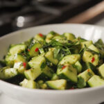 A close-up view of the finished cucumber avocado salad served in a white ceramic bowl on a white marble cooktop. The vibrant green colors of the cucumbers and avocados contrast beautifully with the fresh herbs sprinkled on top, making it ready to enjoy.