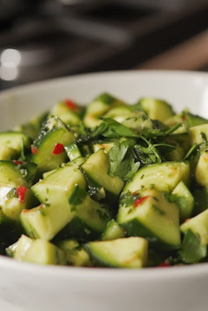 A close-up view of the finished cucumber avocado salad served in a white ceramic bowl on a white marble cooktop. The vibrant green colors of the cucumbers and avocados contrast beautifully with the fresh herbs sprinkled on top, making it ready to enjoy.