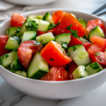 A close-up view of the fresh tomato cucumber salad served in a white ceramic bowl on a white marble cooktop. The vibrant red, green, and white colors shine under soft lighting, making it a refreshing dish ready to be enjoyed.