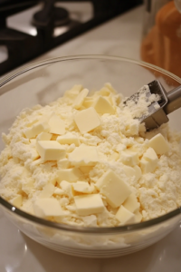 A close-up of a glass mixing bowl on a white marble cooktop, with cold butter pieces being cut into the flour mixture using a pastry cutter. The mixture is turning into coarse, crumbly bits, ensuring a flaky texture for the fritters.