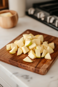 A close-up of a wooden cutting board on a white marble cooktop, displaying freshly peeled, cored, and diced Granny Smith apples. A paring knife rests beside the neatly chopped apple pieces, ready for cooking.