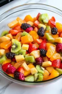 A close-up shot of the same glass mixing bowl on the white marble cooktop, now filled with freshly diced kiwis, strawberries, apple, peach, and mango. The colorful blend of red, green, orange, and yellow fruits creates a visually appealing and vibrant base for the Summer Citrus Fruit Salad.