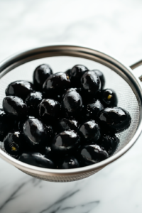 A close-up view of a can of black olives being drained over a small strainer on the white marble cooktop. The olives glisten as they are prepared, ready to be added to the salad for a rich, briny flavor.