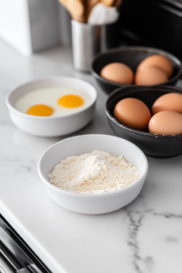 A dredging station set up on the white marble cooktop, consisting of three shallow bowls: one filled with all-purpose flour, another with beaten eggs, and the third with golden panko breadcrumbs. The ingredients are neatly arranged for coating the crab legs.