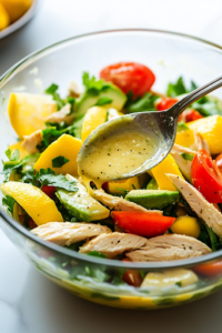 A close-up shot of a glass bowl on a white marble cooktop, as the freshly whisked lemon dressing is poured over the avocado chicken salad. A spoon gently tosses the ingredients, ensuring everything is evenly coated with the tangy dressing.