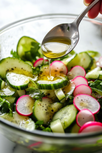 A close-up view of a glass mixing bowl on a white marble cooktop as the prepared dressing is being drizzled over the vibrant salad. A spoon is mid-motion, ready to toss and evenly coat the crisp vegetables with the flavorful dressing.