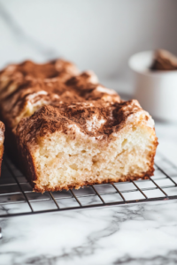 A small glass bowl on the white marble cooktop, where powdered sugar is being whisked with milk to create a smooth glaze. A spoon drizzles the glaze over the cooled cinnamon apple bread, which sits on a wire rack, ready to be enjoyed.
