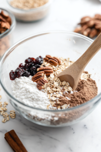 A close-up of another glass mixing bowl on a white marble cooktop, containing whole wheat flour, baking soda, brown sugar, rolled oats, shredded coconut, chopped pecans, dried cherries, cinnamon, salt, and ground ginger. A wooden spoon is partially submerged, ready to combine the ingredients.