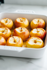 A close-up shot of hollowed apples standing upright in a glass baking dish over the white marble cooktop. A spoon is gently pressing the oat and brown sugar mixture into each apple, ensuring they are evenly filled for a perfect bake.
