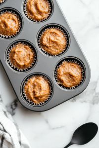 A close-up of a lined muffin tin on a white marble cooktop, with each cup filled about ¾ full of thick, textured batter. A spoon rests beside the tin, with a bit of batter on it, showing the hearty mixture.