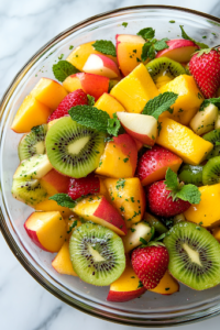 A close-up shot of the fully prepared Summer Citrus Fruit Salad in a clear glass serving bowl on the white marble cooktop. The fresh, glistening fruits are beautifully garnished with finely chopped mint and a drizzle of honey, creating a visually enticing and refreshing dish, ready to be enjoyed.