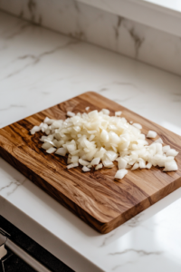 A close-up view of a wooden cutting board on a white marble cooktop with finely chopped onions neatly piled up. The small, even pieces glisten slightly, showcasing their crisp texture and fresh aroma.