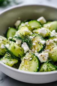 A close-up view of the finished cucumber, avocado, and feta salad served in a white ceramic bowl on a white marble cooktop. Fresh dill and crumbles of feta garnish the dish, adding a final touch of flavor and elegance.