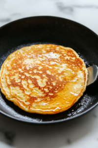 A close-up of a golden-brown pancake being flipped with a spatula on the black skillet over the white marble cooktop. The edges are crisp, and the surface has an even golden color, showing that it's cooked to perfection.