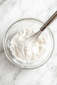 A close-up of a glass bowl on a white marble cooktop, where all-purpose flour and baking powder are being whisked together. A fine dusting of flour coats the edges of the bowl, as the dry ingredients are evenly combined for a smooth cake batter.