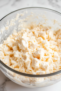 Diced imitation crabmeat being gently folded into the creamy mixture in a glass mixing bowl on the white marble cooktop. The crabmeat is evenly distributed to ensure every bite has a balanced flavor.