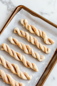 A close-up of a parchment-lined baking sheet on a white marble cooktop, displaying cinnamon-sugar dough strips twisted into neat spirals. The layers of dough and filling are visible, arranged in perfect alignment, ready to bake.
