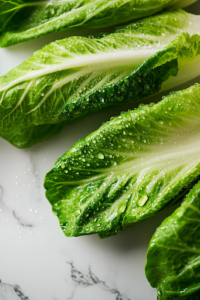 A close-up shot of crisp Romaine lettuce leaves being washed and separated over a white marble cooktop. Tiny water droplets glisten on the fresh green leaves, showcasing their freshness before assembling the wraps.