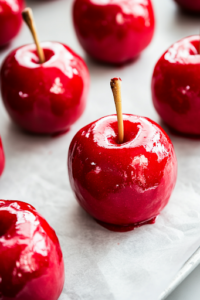 A close-up shot of freshly coated candy apples arranged neatly on a parchment-lined baking sheet over the white marble cooktop. The glossy red candy coating glistens under the light as it begins to harden.