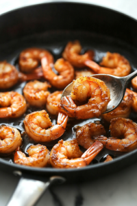 A black skillet on a white marble cooktop filled with hot oil, where coated shrimp sizzle and turn golden brown. A slotted spoon lifts one shrimp, showcasing its crispy, crunchy texture as it finishes frying.
