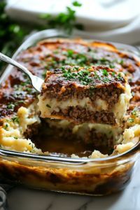 A close-up shot of the fully baked Loaded Mashed Potato Meatloaf Casserole in the glass baking dish on the white marble cooktop. The top is golden and bubbly, garnished with fresh parsley or chives. A serving spatula lifts a slice, revealing the rich layers of meatloaf, mashed potatoes, cheese, and crispy bacon.
