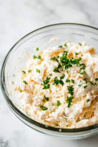 A creamy and well-combined crab dip in a glass mixing bowl on the white marble cooktop, ready to be refrigerated. The dip has a smooth texture with visible bits of crab and green onions.
