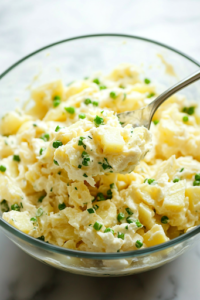 A close-up of the same glass mixing bowl on a white marble cooktop, now containing the fully mixed but chunky potato salad. A spoon is lifting a portion, showcasing the creamy yet textured consistency of the dish.
