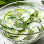 A bowl of chilled German cucumber salad served in a white ceramic dish on the white marble cooktop. The creamy dressing lightly coats the thinly sliced cucumbers, with fresh dill sprinkled on top as a garnish, ready to be enjoyed.