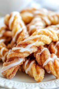 A close-up of warm cinnamon sugar crescent twists elegantly arranged on a white serving plate, drizzled with smooth almond glaze. The glaze glistens under the light, enhancing the golden-brown, flaky pastry layers with an irresistible shine.