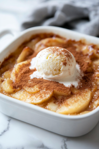 A freshly baked, golden-brown apple dessert resting on the white marble cooktop. The cinnamon-sugar topping glistens, while a scoop of melting vanilla ice cream sits on top, enhancing the warm, spiced flavors.