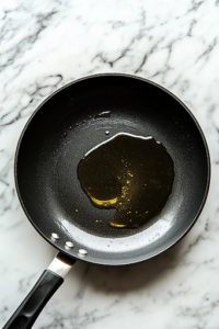 A black frying pan on a white marble cooktop, filled with hot oil heating up to 350°F (175°C). A digital thermometer is submerged in the oil, checking the temperature. The oil shimmers, indicating it is ready for frying.