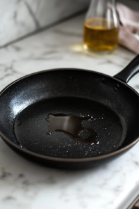 A black skillet on a white marble cooktop with a thin layer of olive oil glistening under the light. The oil is heating up, preparing for the next step of sautéing ingredients.
