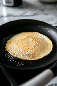A close-up of a black skillet on a white marble cooktop, lightly greased with butter. The pan is heated over medium heat, with a small pat of butter melting on the surface, creating a non-stick layer for perfectly cooked pancakes.