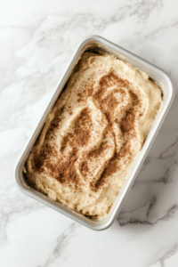 A greased loaf pan on the white marble cooktop, with half of the batter spread evenly inside. A spoon sprinkles a layer of cinnamon-sugar-coated apples over the batter, creating a flavorful filling for the apple fritter bread.