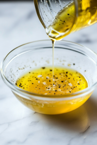 A close-up shot of a small glass bowl on a white marble cooktop, where a zesty dressing of lemon juice, extra virgin olive oil, salt, and pepper is being whisked together. A golden stream of olive oil drizzles into the bowl, blending smoothly.