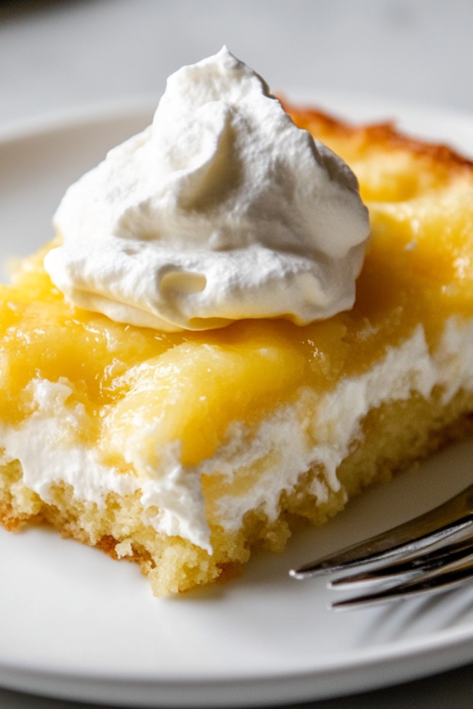 A close-up shot of a plate with a warm slice of lemon cream cheese dump cake, served with a dollop of fluffy whipped topping. The layers of lemon filling, soft cake, and creamy cheese are visible, with a fork ready for the first bite.