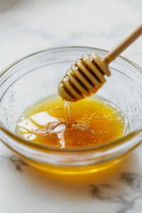 A close-up shot of a small glass bowl placed on the white marble cooktop, where melted butter and golden honey are being whisked together. The mixture has a thick, glossy texture, forming a smooth honey butter glaze.