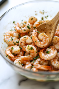 A close-up shot of a glass mixing bowl placed on a white marble cooktop, filled with peeled and deveined shrimp. The shrimp are being tossed with finely minced garlic, olive oil, lime juice, cumin, salt, and black pepper using a wooden spoon. The marinade coats the shrimp evenly, preparing them for cooking.